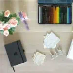 A top-down view of a desk with pink flowers, coloured pencils, the best pocket notebooks, ribbons, cut-out papers with text, three small silver clips, and part of a spiral notebook.