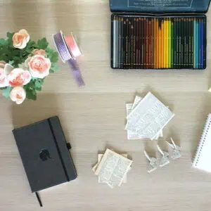 A top-down view of a desk with pink flowers, coloured pencils, the best pocket notebooks, ribbons, cut-out papers with text, three small silver clips, and part of a spiral notebook.