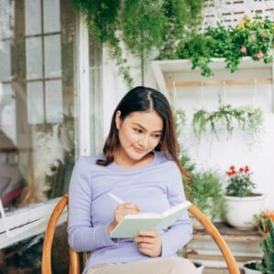 A young woman sitting on a wooden chair writes mindful and reflective journaling exercises in a notebook, smiling softly. She is surrounded by hanging plants and potted greenery, creating a peaceful, garden-like setting near a window.