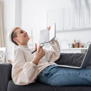 A person sitting on a sofa, smiling and holding up an open notebook featuring a mindful and reflective journal layout, pen in hand, with a laptop on their lap in a bright living room.