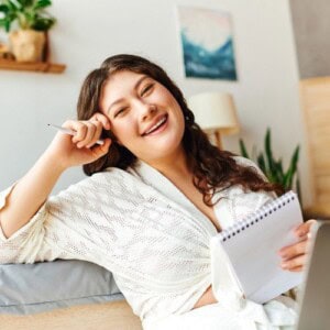 A smiling woman in a white dressing gown sits indoors, holding a pen and a notepad for mindful and reflective journalling, appearing relaxed and cheerful. There are plants and a painting in the background.