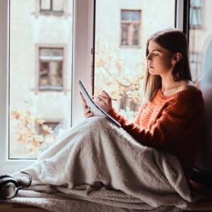 A woman sits by a window wrapped in a blanket, wearing an orange jumper, and writes in a notebook. Natural light fills the cosy scene as she enjoys a more mindful moment, with buildings and trees visible outside.