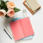 An open pink notebook on a closed blue notebook, a pencil and pen beside them, a crystal paperweight, two closed notebooks in a wooden tray, and a bouquet of light pink roses offer perfect inspiration for your next pocket notebook ideas on this white desk.