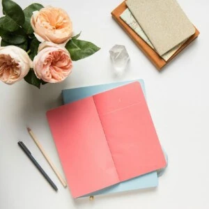An open pink notebook on a closed blue notebook, a pencil and pen beside them, a crystal paperweight, two closed notebooks in a wooden tray, and a bouquet of light pink roses offer perfect inspiration for your next pocket notebook ideas on this white desk.
