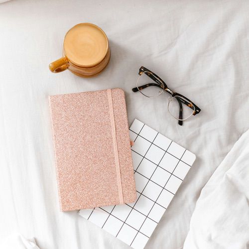Pocket Notebook Layout 2 A flat lay photo featuring a pink glittery pocket notebook layout, a black-and-white grid-patterned notepad, tortoiseshell glasses, and a mug of coffee on light-coloured bed sheets.