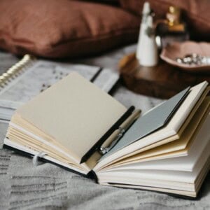 An open pocket notebook layout with blank pages and a pen resting inside sits on a bedspread, with a diary, pillows, and a tray holding jewellery and small items in the background.
