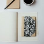 An open lined notebook with a small, closed, black-and-white patterned pocket notebook on top creates a perfect pocket notebook setup next to a pencil, notepad, and coffee cup on a white surface.