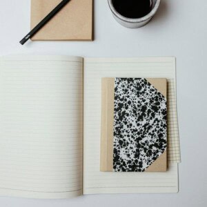 An open lined notebook with a small, closed, black-and-white patterned pocket notebook on top creates a perfect pocket notebook setup next to a pencil, notepad, and coffee cup on a white surface.