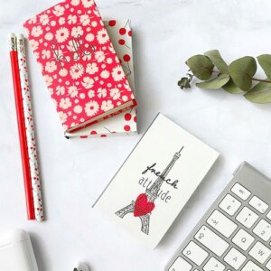 Two patterned exercise books, two red-and-white pencils, a small pocket notebook featuring the eiffel tower and “french attitude” text—perfect for exploring pocket notebook uses—eucalyptus leaves, wireless earphones case, and part of a white keyboard on a white surface.