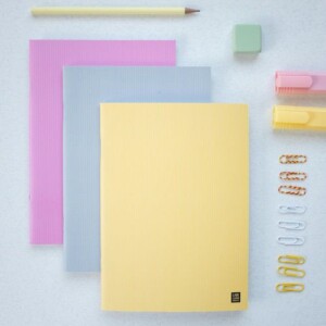 This series of pocket notebooks features pastel covers in yellow, grey, and pink, arranged on a light surface with a yellow pencil, green eraser, two highlighters, and sets of paperclips in yellow, white, and gold.