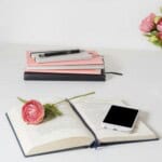 An open book with a pink flower and a mobile phone rests on a white desk. In the background, stacked notebooks hint at things to write in a pocket notebook, while a vase with pink flowers decorates the side.