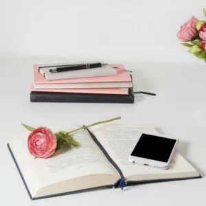 An open book with a pink flower and a mobile phone rests on a white desk. In the background, stacked notebooks hint at things to write in a pocket notebook, while a vase with pink flowers decorates the side.