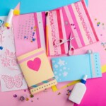 Colourful craft supplies and handmade cards decorated with hearts, stars, and glitter are arranged on a pink and blue background—perfect materials to customise a pocket notebook. Scissors, glue, stickers, and stencils are also visible.
