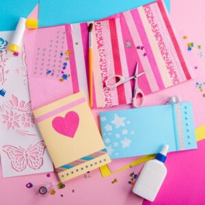 Colourful craft supplies and handmade cards decorated with hearts, stars, and glitter are arranged on a pink and blue background—perfect materials to customise a pocket notebook. Scissors, glue, stickers, and stencils are also visible.
