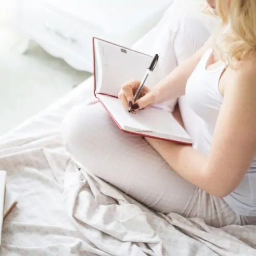 A woman in light-coloured pyjamas sits cross-legged on a bed, journalling for improved sleep in an open notebook with a black pen. The scene is bright and peaceful, with soft bedding and natural light.