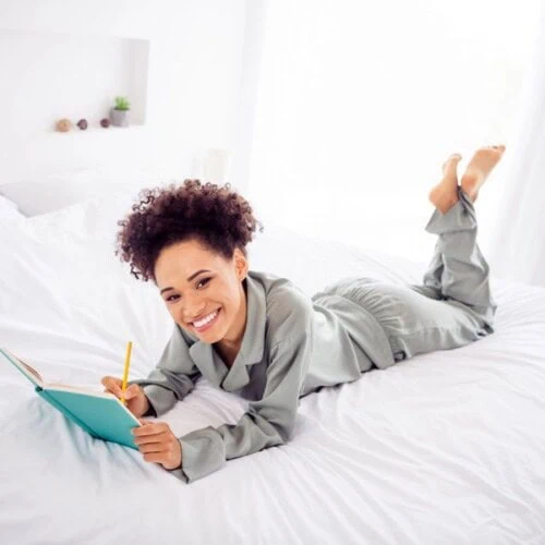 A smiling woman in pyjamas lies on her stomach on a white bed, journalling for improved sleep in a teal notebook with a yellow pencil. The room is bright with natural light and features minimal, cosy decor.