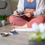 A person sits cross-legged on the floor, journalling for stress management. Wearing a blue sports bra, light jumper, and pink trousers, they’re surrounded by plants, crystals, and a pack of cards on a light wooden floor.