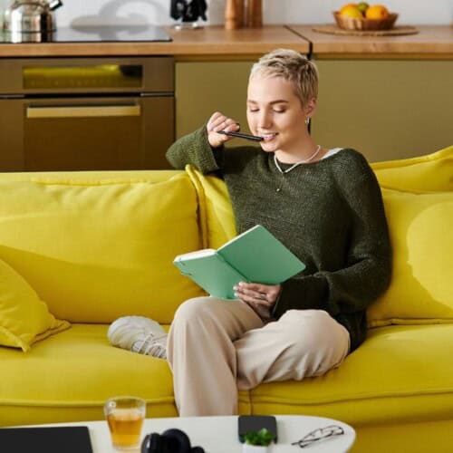 A person with short blond hair sits on a yellow sofa, smiling and holding a mint-green notebook while lightly biting a pen—perhaps journalling for stress management. A mug and tech devices are on the table, with a kitchen in the background.