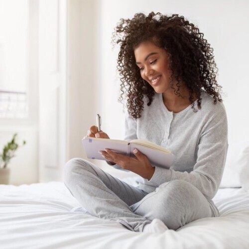 A woman with curly hair, dressed in light grey pyjamas, sits cross-legged on a bed and smiles while writing in a notebook—embracing nightly journalling to improve sleep. The room is bright and cosy with natural light streaming in.