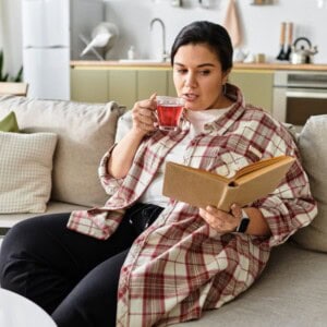 A person sits on a sofa in a cosy living room, sipping tea from a glass mug and reading a book filled with stress relief journal prompts. The background reveals a kitchen area with appliances and shelves.
