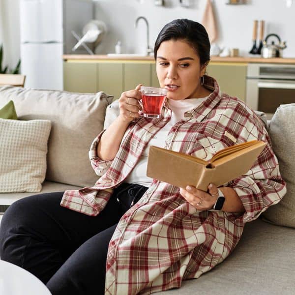A person sits on a sofa in a cosy living room, sipping tea from a glass mug and reading a book filled with stress relief journal prompts. The background reveals a kitchen area with appliances and shelves.