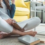 A person practising stress relief journalling techniques sits cross-legged on a rug, holding a notebook and pen, with books nearby. A yellow sofa and white table are in the background.