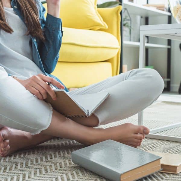 A person practising stress relief journalling techniques sits cross-legged on a rug, holding a notebook and pen, with books nearby. A yellow sofa and white table are in the background.