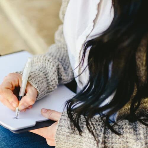 Close-up of a person with long dark hair in a knitted jumper, writing in a notebook with a pen—practising stress relief journalling techniques. The focus is on the hand and the notebook.