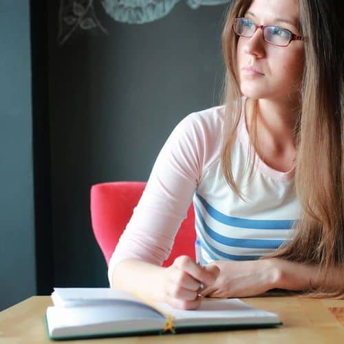 A woman with long brown hair and glasses sits at a desk, holding a pen over an open notebook, looking thoughtfully to the side as she considers her next entry among the grief journal prompts.