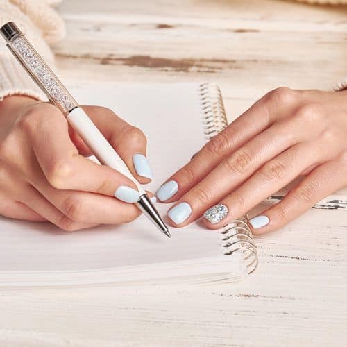 A person with light blue painted nails writes grief journal prompts in a spiral notebook with a decorative pen on a white wooden table.