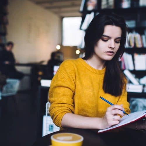 A woman in a yellow jumper writes in a notebook at a table in a cosy, modern café with bookshelves in the background, thoughtfully exploring grief journal prompts. A cup of coffee sits in the foreground.