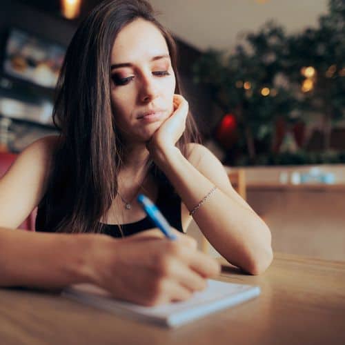 A woman sits at a table, resting her chin on one hand whilst writing grief journal prompts in a notebook with a pen. She looks thoughtful and focused, with a blurred background of indoor plants and soft lighting.