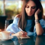 A woman with long brown hair writes in a small notebook at a table with a coffee cup, wearing a blue blouse and silver bracelets, looking thoughtful as she reflects on her emotions through grief journalling.