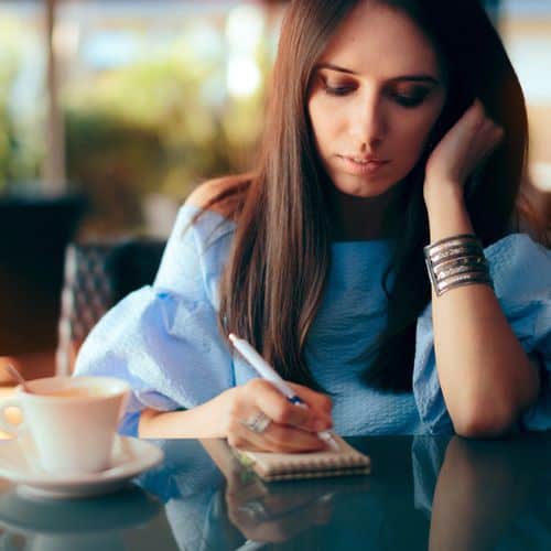A woman with long brown hair writes in a small notebook at a table with a coffee cup, wearing a blue blouse and silver bracelets, looking thoughtful as she reflects on her emotions through grief journalling.