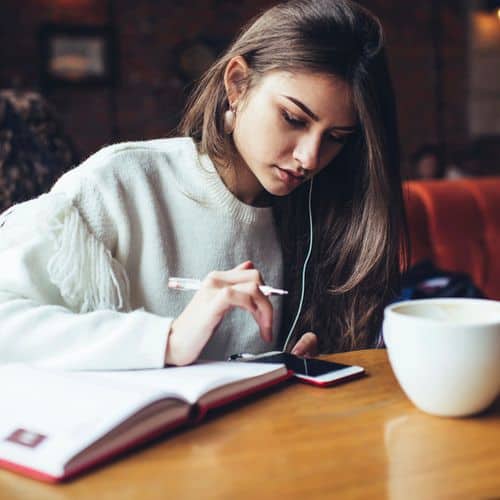 Young woman with long hair sits at a table, writing in a notebook whilst looking at her phone. She wears a white jumper, has headphones in, and seems focused on grief journalling. A large mug is on the table next to her.