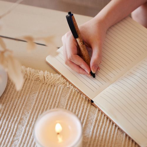 A hand writes in a lined notebook on a beige table next to a lit candle and some dried plants, creating a peaceful space for grief journalling.