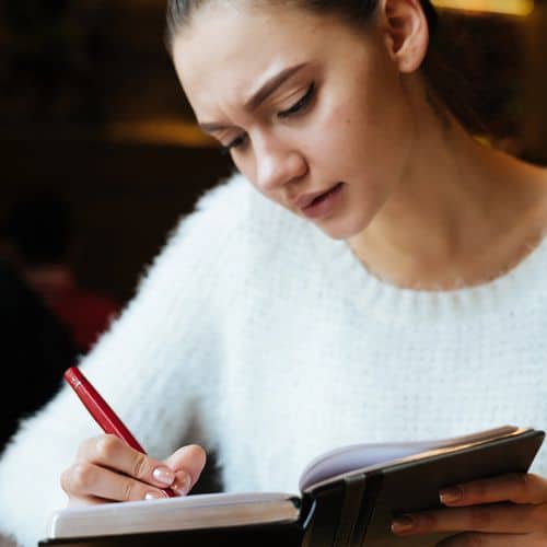 Young woman with brown hair in a white jumper writes with a red pen in a notebook, appearing focused and thoughtful as she engages in grief journalling exercises.