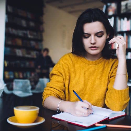 A woman in a yellow jumper sits at a table in a café, writing grief journalling exercises in her notebook with a cup of coffee beside her. Bookshelves are visible in the background.