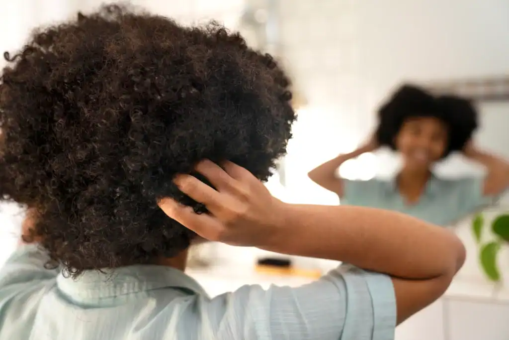 Young black woman touching her hair and looking in the mirror. Deep Conditioners For Curly Hair