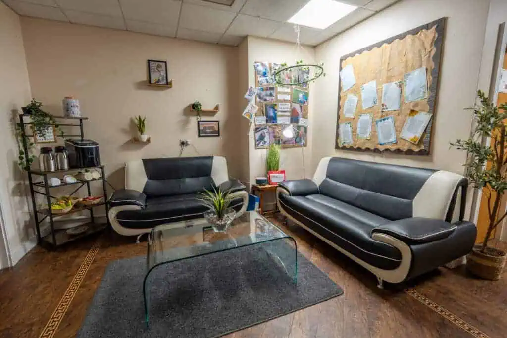 Comfortable nursery waiting room with stylish black and white sofas, glass coffee table, and decorative plants, creating a warm and inviting space for parents and visitors.
