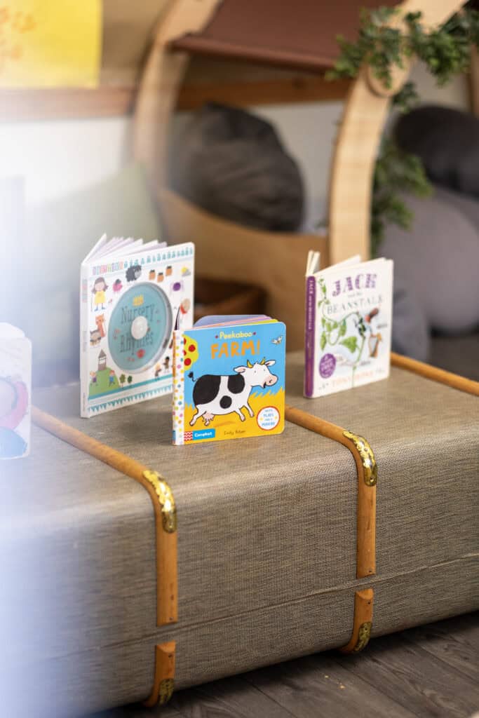 Bright children's books about farming, nursery rhymes, and nature on a beige storage chest in a colourful playroom, promoting early childhood learning at Thrive Childcare.