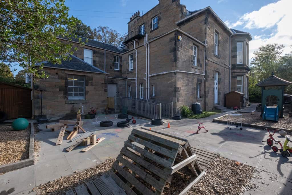 Bright outdoor playground at Thrive Childcare with toys, ride-on bikes, and play equipment on a paved area beside a historic stone building, providing a safe and engaging environment for young children.