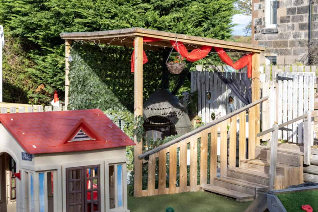 A colourful outdoor play area at Thrive Childcare with a cozy rattan nest, wooden fencing, and festive red decorations, promoting safe, engaging, and creative childcare environments.