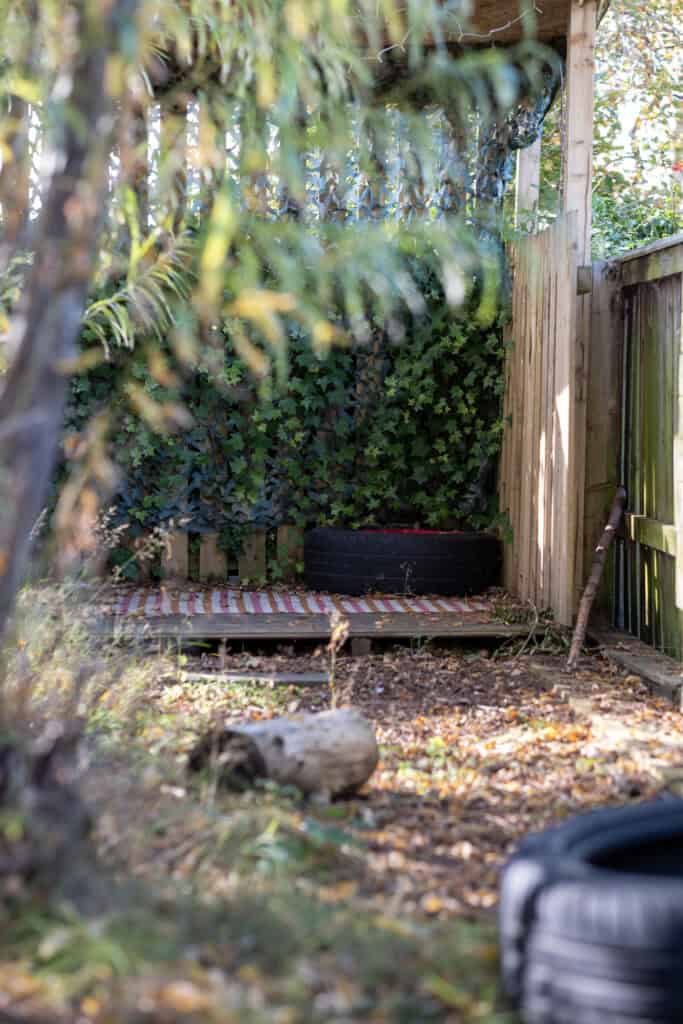 A small outdoor play area with wooden fencing, surrounded by greenery and ivy, featuring a tire and wooden platform for children's outdoor activities at Thrive Childcare.