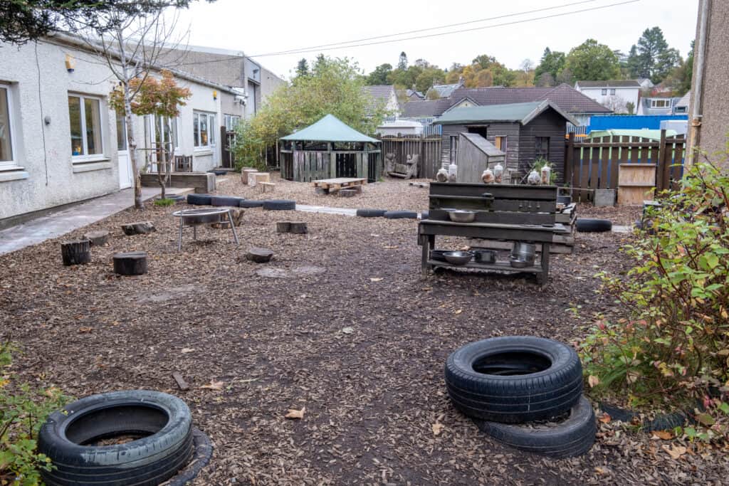 A spacious outdoor play area at Thrive Childcare, featuring wooden logs, tires, and natural elements, designed for outdoor activities and early childhood development. Perfect for community and learning.