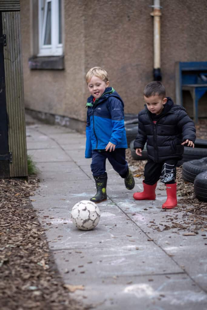 1. Two young children playing football outdoors at Thrive Childcare, dressed warmly in jackets and boots, enjoying active outdoor play and socialising with friends.