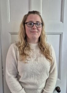 1. A woman with glasses and long curly hair standing in front of a white door, representing professional staff at Thrive Childcare in the UK.