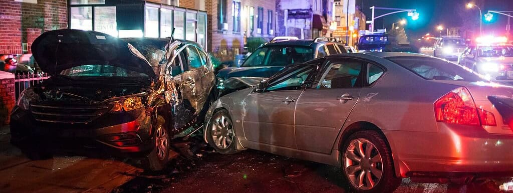 Black car involved in a multi-vehicle crash with visible front-end damage at night, police vehicles with flashing lights in the background, city street scene after an accident, emergency response.