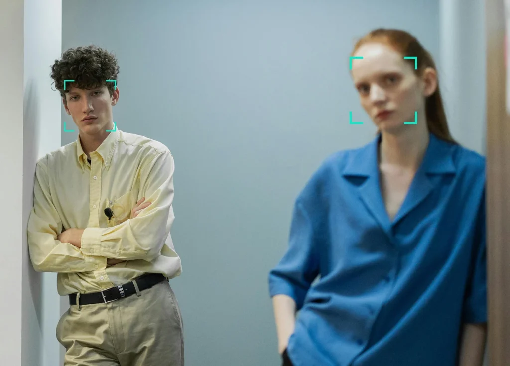 Two people stand in a hallway, one in a yellow shirt with arms crossed, the other in a blue shirt. Both have green facial recognition squares around their faces.