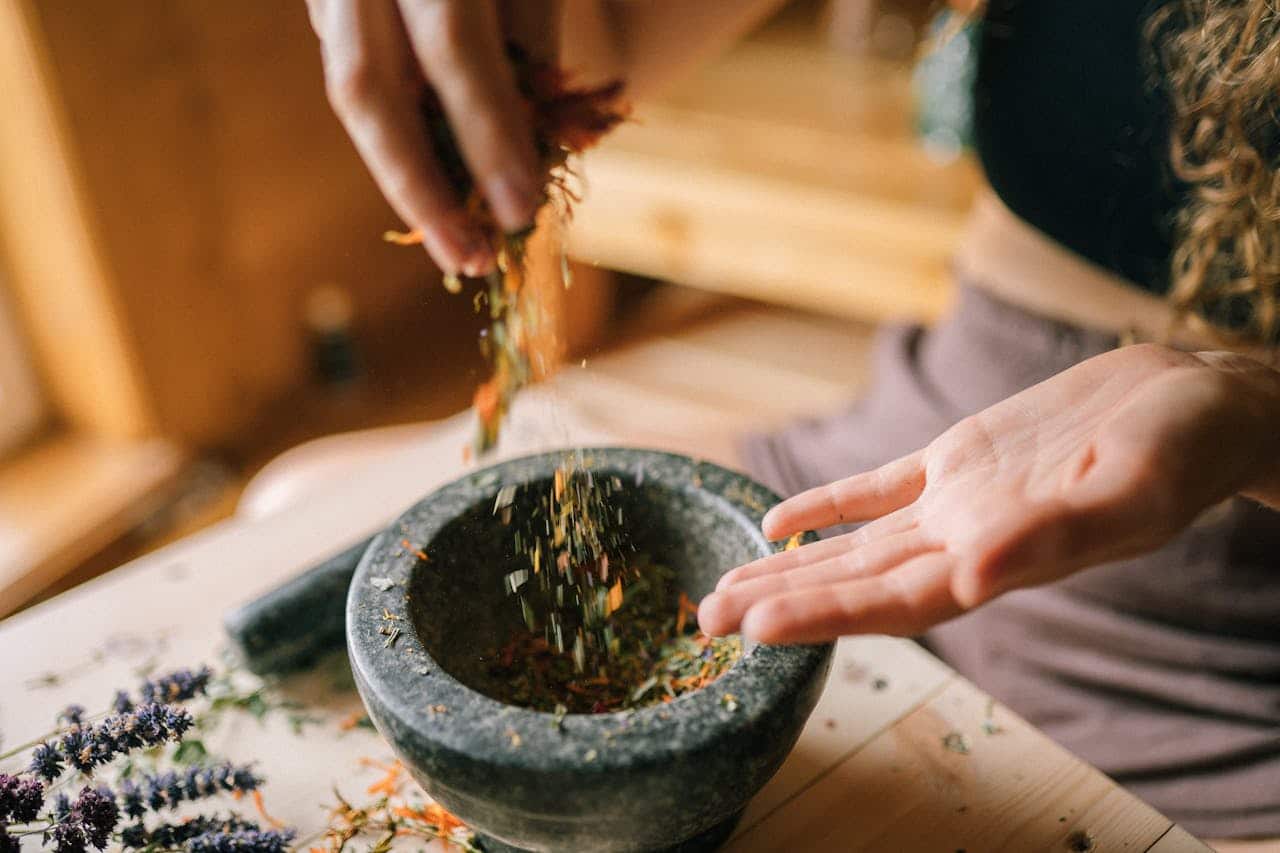A person preparing dried herbs using a mortar and pestle on a wooden table, seen from above.